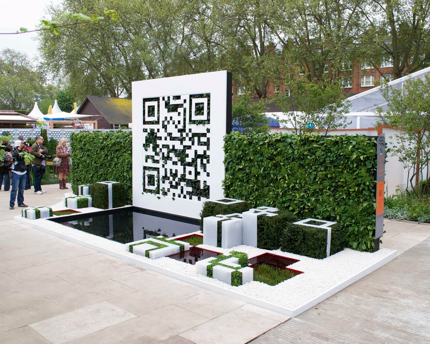 A large outdoor QR code made from Re-Board in front of a leafy green garden display.