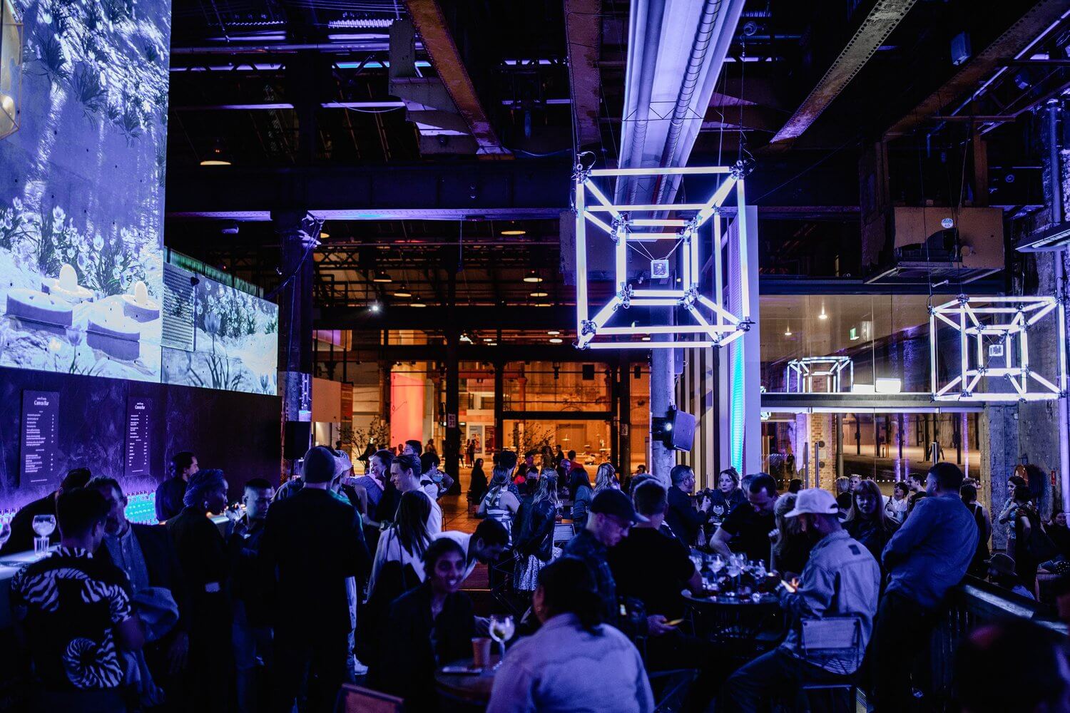 A large group of people in a dimly lit warehouse looking bar with large neon cubical installations and screens around the room.